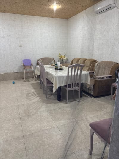 The image shows an indoor scene, likely a dining or living area, within a residential setting in Serebryakovka, Russia. The room features tiled flooring, a light-colored textured wallpaper on the walls, and a wood-paneled ceiling with an overhead light fixture. An air conditioning unit is mounted high on one wall.
In the center of the room is a dining table covered with a white tablecloth. Several chairs, upholstered in a light purple or pinkish fabric, are arranged around the table. Some of these chairs appear to be a light gray or lavender color. On the table, there are various items, including what appear to be food containers, a glass dome, and a vase with a floral arrangement.
To the left of the dining area, a child's high chair with a purple seat is positioned near the wall. A small blue ball is visible on the floor nearby. In the background, a sofa and armchairs, upholstered in brown fabric with patterned throws, are visible. One armchair has wooden armrests. A glass cloche with items underneath is also on the table, along with jars and bottles.
To the right of the scene, part of another chair with a pinkish upholstered seat is visible, suggesting further seating arrangements or a separate seating area. The overall impression is of a functional, lived-in space.