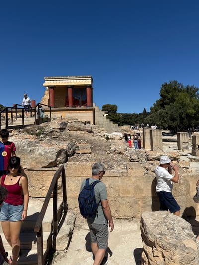The image depicts a sunny outdoor scene at the Palace of Knossos, a Minoan archaeological site located south of Heraklion in Crete, Greece. Several tourists are visible, some walking on stone pathways and stairs, while others observe the ruins. In the foreground, a woman in a red tank top and denim shorts is walking down stairs, while a man with a backpack walks past her. In the mid-ground, a man in a white t-shirt and hat drinks from a bottle. Behind them, ancient stone structures, including pillars and walls, are evident, with a portion of a reconstructed building featuring red columns and a fresco. The background shows more ruins and trees under a clear blue sky. The site appears well-maintained with pathways and railings for visitors.