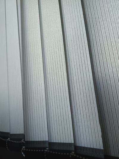 The image displays a close-up, angled view of a set of vertical blinds. The blinds are a light grey color and have a textured, ribbed surface. They are partially drawn, revealing multiple overlapping slats that create a linear pattern. The bottom edge of the blinds is visible, showing a darker hem and a chain of small, white beads, which are part of the blind's operating mechanism. The lighting suggests an indoor setting, possibly near a window, casting subtle shadows that enhance the texture of the blinds. The scene is devoid of any people or specific environmental markers that would indicate a location within Kalmykivka, Ukraine.