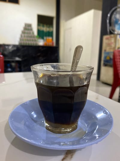 A close-up, eye-level, medium shot captures a glass of dark coffee with a spoon inside, resting on a light blue saucer. The coffee fills about two-thirds of the glass, with a lighter brown layer at the bottom, possibly indicating sugar or creamer. The glass has a faceted design and condensation on its exterior. The saucer is slightly wet, with a few drips of coffee. The background is blurred, showing a white table, and further back, a stack of cans or containers forming a pyramid shape. To the right, a blurry red chair and a refrigerator-like appliance are visible. The scene appears to be indoors, possibly in a cafe or restaurant in Luengbaro, Indonesia, given the context. There are no people visible in the image.
