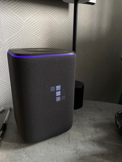 A close-up shot shows a dark gray speaker with a thin blue light strip on its left edge. It's mounted on a black arm extending from a light-colored patterned wall to the left. Behind the speaker, there is a gray countertop or table. A small black cylindrical object is partially visible behind the speaker. At the bottom right corner, a pen is visible. The location is not discernible from the image.