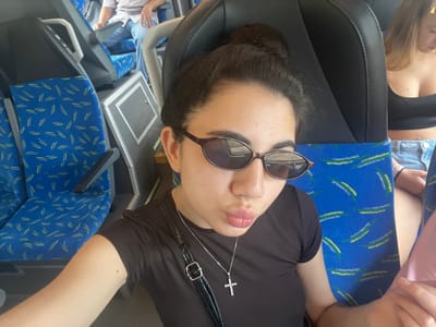 In the foreground, a young woman with her hair in a bun and wearing a dark brown t-shirt, sunglasses, and a silver cross necklace is looking at the camera and puckering her lips. She is seated on a bus with blue seats. To her right, another woman wearing a black top and denim shorts is visible. In the background, a person wearing a light-colored patterned shirt and blue jeans is seated on a bus seat. The environment appears to be the interior of a bus, possibly in Vejprnice, Czechia.