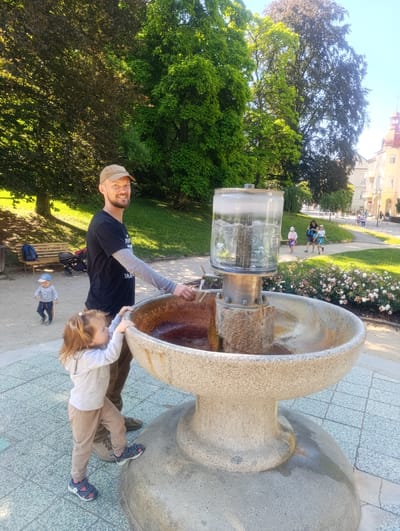 In Luhačovice, Czechia, a man wearing a beige baseball cap and a black t-shirt with a long-sleeved grey undershirt stands by a large, circular stone fountain filled with reddish-brown water. A young girl with blonde hair pulled into a ponytail, wearing a light grey hooded sweatshirt and beige pants, stands next to him, reaching towards the fountain. The man's right hand is near the fountain's edge, and the girl's left hand is touching the rim. In the background, a young boy in a striped shirt and dark pants walks away from the fountain, heading towards a park area. A wooden bench and a stroller are visible further back. Lush green trees and a clear blue sky dominate the upper part of the image, with a few buildings and people visible in the distance along a paved walkway lined with blooming pink flowers. The ground around the fountain is paved with light-colored square tiles.