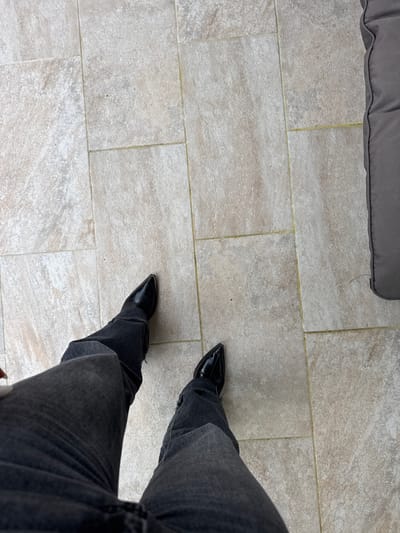 A high-angle shot shows a person standing on a tiled surface. The person is wearing dark-gray jeans and black pointed-toe boots. A gray cushion is partially visible in the upper-left corner. The location is not discernable from this image, therefore Les Arcs, France cannot be determined.