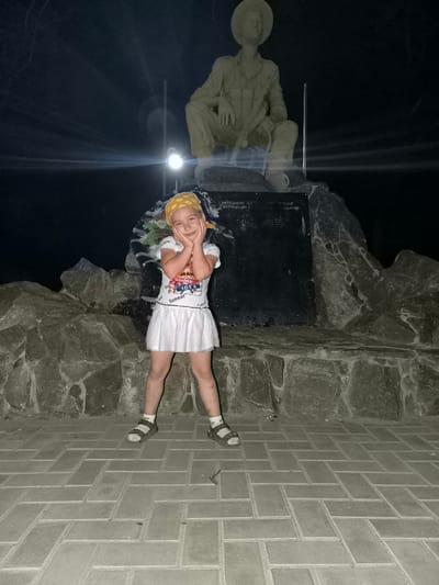 A young girl with a yellow bandana and a white t-shirt with a graphic print and a white skirt stands in front of a monument at night in Vulcănești, Moldova. She is posed with her hands on her cheeks, looking at the camera. The monument appears to be a statue of a soldier sitting on rocks. The ground in front of the monument is paved with rectangular bricks. The scene is lit by artificial lights.