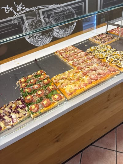 The image captures a selection of rectangular pizzas displayed behind glass in what appears to be a food establishment, likely a pizzeria, in Villafranca di Verona, Italy. The pizzas exhibit various toppings, including salami, vegetables (possibly zucchini and peppers), ham, and some that have a combination of purple cabbage, mushrooms, and cheese. The display case rests on a light wooden surface. A portion of the tiled flooring, in reddish-brown tones, is visible in the upper-right corner. The reflection in the glass reveals a black background with white botanical illustrations.