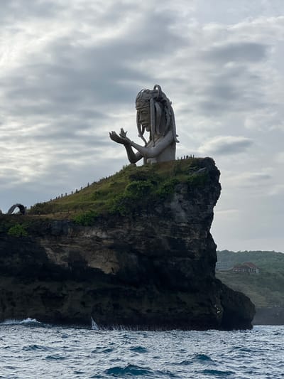The image displays a monumental stone statue situated on a prominent cliff overlooking a body of water. The location is specified as the city of Sakti, Indonesia.

The central subject is a colossal light-colored statue depicting the upper torso of a human-like figure. The figure wears an elaborate headdress, features a defined face, and has numerous thick, rope-like or serpentine elements forming its hair or dreadlocks. Its arms are raised with hands open, palms facing upwards, fingers slightly curled. The statue appears to emerge from the peak of a grassy, rocky promontory.

Small human figures are visible near the base of the statue on a narrow, fenced pathway, providing a sense of scale. Further along the cliff edge to the left, a decorative arched structure is present. The promontory forms a dark, rugged cliff face that descends directly into the choppy, dark blue-green ocean below, where white foam indicates wave action against the rock. In the distant background on the right, a building with a reddish-brown roof and light walls is visible on a lower, green-covered hill. The sky is overcast with a blanket of grey and white clouds. The perspective is from the water, looking upwards at the landmass and statue.