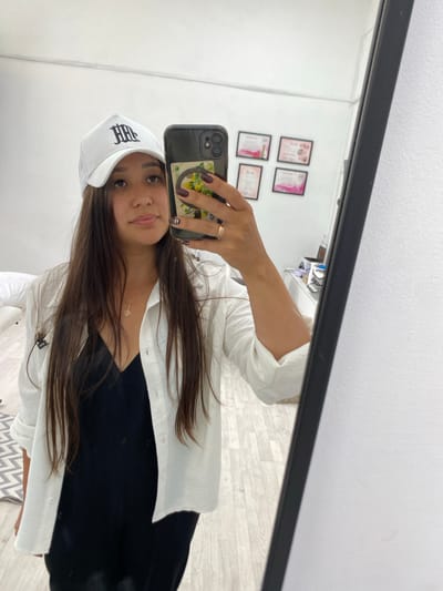 A young woman with long, dark hair is taking a mirror selfie in what appears to be a room with light-colored walls and wooden flooring. She is wearing a white baseball cap with a black logo, a white unbuttoned shirt over a black dress, and a gold necklace. She is holding a smartphone in her right hand, which has a phone case with a floral design. A ring is visible on her right ring finger. In the background, several framed certificates are hanging on the wall, and a mirror reflects the room. The overall setting suggests an indoor, possibly domestic or commercial, environment. The location is described as Akyar, Russia.