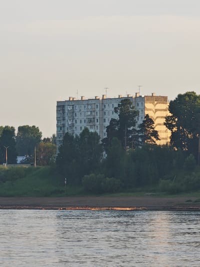A multi-story apartment building with a gray facade and numerous windows stands amidst trees. The building's right side features a distinctive pattern of curved yellow and gray shapes. A body of water, likely a river, occupies the foreground, reflecting the sky and distant lights. A grassy embankment separates the water from the apartment building and dense green trees. The sky is a pale, hazy yellow, suggesting late afternoon or early morning light. The scene appears to be set in Lesosibirsk, Russia, as indicated by the architectural style and surrounding vegetation. There are no people or distinct objects visible on the bank or in the water.