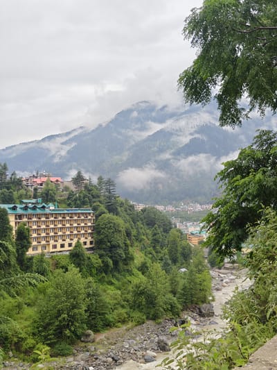 In the city of Aleo, India, a resort-style building with a yellow facade and green-tiled roof is situated on a lush green hillside. This large building has many windows and balconies. Behind it, further up the slope, are several other buildings, some with red roofs and others with green roofs, suggesting a developed area within the mountainous terrain. The mountains in the background are partially obscured by mist and clouds, with trees covering their lower slopes. A winding river, with rocky banks and light-colored water, flows through the valley in the foreground. Dense green foliage from trees and bushes frames the scene, with a large tree with dark green leaves partially obscuring the view in the upper right corner. The overall atmosphere is that of a natural landscape with significant human development, likely a tourist destination, under overcast skies.