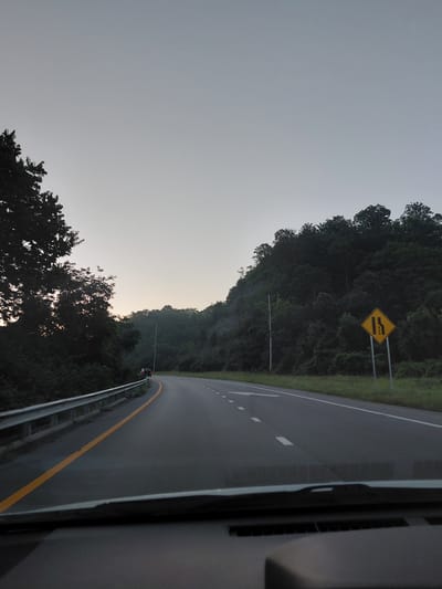 The image captures a view from a vehicle driving on a highway towards a forested hill. The sky is overcast and a pale gray, indicating early morning or late evening. On the left side of the road, a guardrail runs along the shoulder, bordered by a yellow line. Dense trees line the embankment to the left. On the right side of the highway, a yellow diamond-shaped sign with black markings indicates a lane reduction. Beyond the sign, the highway curves around a steep, tree-covered hill. A few cars are visible on the road ahead, some pulled over on the shoulder near the guardrail. The overall scene suggests a rural highway setting.