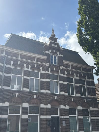 The image shows the exterior of a brick building with white accents and a dark tiled roof. The building has multiple windows with white frames. Above some of the windows, there are decorative brickwork arches. The year "1908" is visible in a stone plaque on the upper facade of the building. The street name "STATIONSSTRAAT" is inscribed above a window on the right side of the building. The building is located in Boxtel, Netherlands. A large green tree is visible on the right side of the image, and the sky above is blue with scattered white clouds. The image is taken from a low angle, looking up at the building. The house number "87" is visible near the bottom of the building.