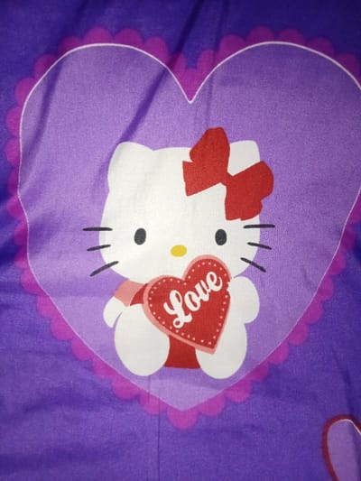 A close-up photograph depicts a graphic design on a purple textile, located in Umsobomvu Local Municipality, South Africa. The central subject is a white cartoon character, styled as Hello Kitty, with black eyes, a yellow oval nose, and three black whiskers on each side of its face. A red bow is positioned on the character's upper left ear. The character is depicted holding a red heart, which has a white scalloped border and the word "Love" written in white script across its center. The character and the heart it holds are contained within a larger, translucent purple heart shape, which is outlined by a scalloped magenta border. The background is a solid dark purple fabric, exhibiting minor wrinkles.
