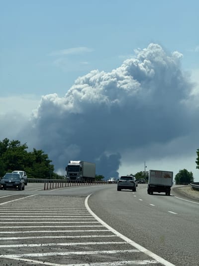 The image shows a highway with several vehicles, including cars and trucks. In the background, a large plume of smoke rises into the sky, obscuring the sun and casting a shadow over the scene. The smoke appears to be coming from a fire in the distance. The sky is partly cloudy. The overall impression is one of a developing weather event or a significant industrial incident. There is no information in the image to suggest the location is in the city of Рождественская, Russia.