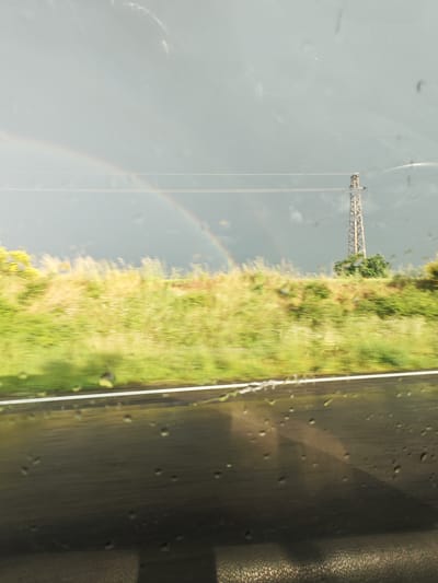 The image shows a view from inside a vehicle, likely a car, with raindrops visible on the window. The foreground features a blurred road and dashboard. Beyond the road, there's a strip of green vegetation. In the background, a faint rainbow is visible in a cloudy sky. An electrical transmission tower stands amidst the foliage. The location is suggested to be near Bogdanovo, Bulgaria, based on the prompt. The subjects include the landscape, weather phenomenon (rainbow), and infrastructure (transmission tower). The action is the viewer observing this scene from a moving vehicle.