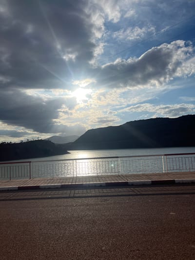 The image shows a landscape, likely taken in Tabaroucht, Morocco. The view includes a body of water reflecting sunlight, a mountain ridge silhouetted against a partly cloudy sky, and a roadside or walkway. The sun is visible through the clouds, creating a lens flare effect. A roadside structure or barrier with a grid-like design is in the foreground. The pavement appears textured.