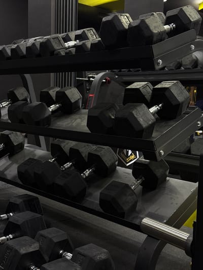 A weight rack filled with black, hexagonal dumbbells. The rack is made of dark metal and appears to have a multi-tiered design. The weights are arranged in rows, showcasing various sizes. The lighting casts shadows, emphasizing the texture and form of the equipment. The weights include markings such as "17.5 kg" and "27.5 kg". A sticker is placed near the weights, and an element made of silver metal, potentially a handle, is placed at the right of the frame.