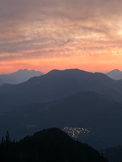 The image captures a panoramic view of a mountainous landscape at dusk. In the foreground, the silhouette of a dark, forested mountain ridge dominates the lower portion of the frame, with a few scattered trees visible on its left side. In the mid-ground, a cluster of lights, presumably from a village or town, twinkle in the valley between mountain ranges. Further into the distance, several layers of mountain peaks are visible, fading into the atmospheric haze. The sky above is painted with the warm hues of a sunset, transitioning from a soft orange glow near the horizon to shades of pink and purple, with subtle cloud formations scattered across it. The scene suggests a remote, tranquil setting as day gives way to night. The village lights are likely located in or around San Juan de Plan/San Chuan de Plan, Spain, a municipality in the province of Huesca, within the autonomous community of Aragon.