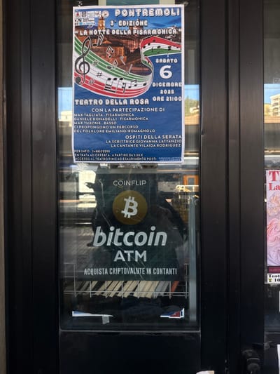 This image depicts a close-up, frontal view of a glass pane, likely a storefront window or door, in the city of La Spezia, Italy. Affixed to the upper section of the glass is a rectangular blue poster advertising "Pontremoli 3° Edizione La Notte della Fisarmonica" (The Night of the Accordion), scheduled for Saturday, December 6, 2025, at 9:00 PM at the Teatro della Rosa. The poster features a large treble clef stylized with red, white, and green Italian flag colors and musical notes, overlaid on a background image of a European townscape. Details include participating accordionists Max Tagliata and Daniele Donadelli, bassist Max Turone, and guest performers Giovanna Lattanzio and Vilaida Rodriguez, presenting Emilian/Romagnolo folklore. Entry is by offer starting from €5.00.
Centered on the lower half of the glass is a "Coinflip Bitcoin ATM" advertisement, featuring a prominent yellow Bitcoin 'B' logo on a dark background. Text states "ACQUISTA CRIPTOVALUTE IN CONTANTI" (Buy cryptocurrencies with cash). Both the poster and the Bitcoin ATM advertisement show minor creasing and wear.
The glass pane reflects elements of the surrounding environment, including an indistinct partial reflection of the photographer and lighter areas indicative of sky or distant buildings. Behind the glass, visible below the Bitcoin ATM sign, are dark, stacked items, possibly magazines or books. To the right, another partial poster with light pink graphics and "Teatro" text is faintly visible through the glass. The glass is framed by dark, vertical mullions or door frames. The scene is illuminated by ambient daylight.