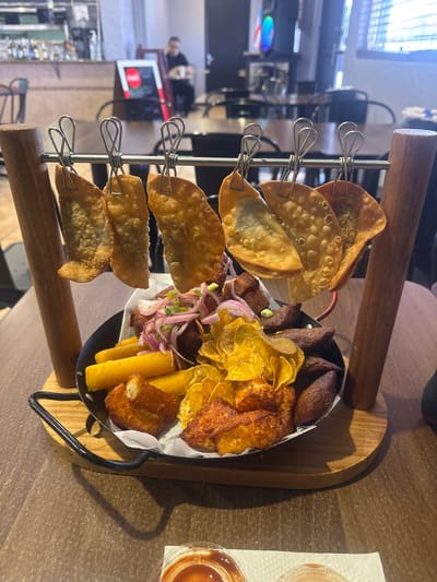 This is a medium shot, eye-level, close-up photograph of a food platter. The platter is round, black, and sits on a wooden base, which is on a wooden table. The platter contains various fried foods, including what appear to be empanadas hanging on clips above the platter. Below the empanadas is a mound of fried plantain chips, yuca fries, and what seem to be small, fried meat items. There are also pieces of fried cheese or dough, and some thinly sliced red onions and cilantro. In the background, blurred, is a restaurant interior with tables, chairs, and a counter with bottles and what might be a menu board. The lighting is natural and artificial.