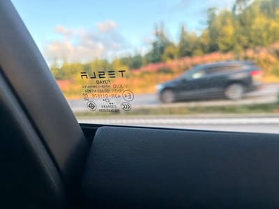The image shows a close-up view from inside a vehicle, looking out the side window. The window has various markings and text etched into it. The prominent marking is the "TESLA" logo. Below the logo, there is text that appears to be a safety or manufacturing code, including characters like "OAYU", "V-X1ID", and numbers that are partially obscured or difficult to read due to reflection and focus. There are also circular symbols with additional markings within them, some resembling certifications.

Through the window, a blurred view of a rural landscape is visible, with trees and vegetation in the background. A dark-colored vehicle, possibly an SUV, is seen driving on a road in the distance, also blurred. The sky is visible, showing a light blue color with some clouds.

The foreground of the image shows the interior of the car, specifically the black plastic trim of the door panel and a portion of the seat or armrest. The lighting suggests it is daytime, likely afternoon or evening, given the quality of light and the sky color. The scene is captured from a stationary or slow-moving vehicle.

The image does not provide any specific visual cues or landmarks that would definitively place it in Munkedal, Sweden. Without additional context or identifying features in the visual scene itself, it is not possible to confirm the location.