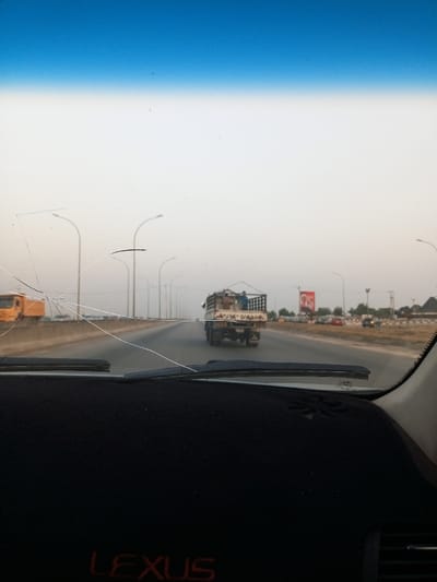 The image is a view from the driver's seat of a car. The windshield has a large crack radiating from the top left corner to the bottom right corner of the image. The car is traveling down a four-lane road in Dushekoro, Nigeria (9.128, 7.369). There are streetlights lining the sides of the road. A white flatbed truck is traveling in the lane to the right of the car. There are other vehicles in the distance. The sky is blue with a layer of white haze. The car's dashboard is visible in the foreground. The dashboard is black with the word "LEXUS" written in red across the bottom of the image. The windshield wiper is in the down position.