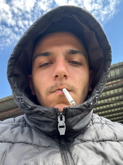 A young man wearing a black puffer jacket with the hood up is looking downward into the camera. He has a cigarette in his mouth. The background shows a blue sky with scattered clouds and the edge of a metal structure. The image appears to have been taken outdoors. No information is present to confirm this took place in Srednia, Bulgaria.
