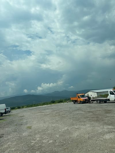 This image depicts an outdoor, unpaved gravel lot located in Karnare, Bulgaria, under an overcast sky. The horizon reveals multiple layers of mountains extending into the distance, appearing progressively lighter in tone due to atmospheric perspective. In the mid-ground, a simple, white, single-story building with a light-colored roof is situated on the right side. Utility poles and power lines are visible in the landscape between the building and the mountains.
The gravel lot itself shows sparse patches of green vegetation. On the far left, a portion of a white panel van is parked, with a silver hatchback visible behind it. Closer to the center, an orange dump truck is parked with its driver's side door open. This truck features a distinctive red and white striped reflective decal across its front bumper/grille. Adjacent to the orange truck, a white flatbed tow truck is positioned, pulling a trailer that is loaded with a dark, granular material, likely gravel or soil.
Two male individuals are present in the scene. One man, dressed in a dark shirt and light trousers, is seen bending down near the open door of the orange dump truck, appearing to be engaged in an activity involving the vehicle. The second man, wearing a white t-shirt and dark trousers, stands upright between the orange truck and the white flatbed truck, observing the activity.