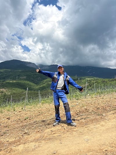 A full-body, vertical, medium-shot photograph captures a person standing in a vineyard with a mountainous backdrop under a cloudy sky. The person, who appears to be female and is smiling, is wearing a blue work uniform with grey accents and a white baseball cap. Her arms are extended outwards, and her body is angled slightly to the viewer's left. The vineyard is characterized by rows of young plants supported by metal stakes and wires, situated on a dirt path. In the background, lush green mountains rise, with some rocky outcrops visible. The sky is dominated by dark, heavy clouds, with patches of blue sky interspersed. The overall impression is of an outdoor, agricultural setting.