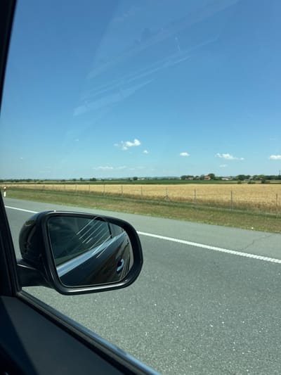 The image shows a view from inside a car traveling on a highway. A side-view mirror is visible in the foreground, reflecting the sky and the car's interior. Beyond the mirror, a golden field stretches to the horizon, bordered by a line of trees and houses. The sky is clear and blue with a few scattered white clouds. A white line marks the edge of the highway. This scene is consistent with the rural landscape found in Indjija Municipality, Serbia.