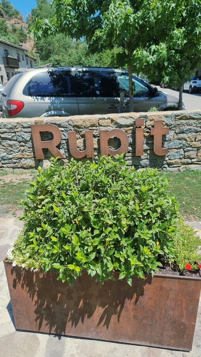 The image shows a sign with the word "Rupit" made of rusty metal letters attached to a stone wall. In front of the sign is a large planter box filled with lush green bushes and some small white flowers. To the left and behind the bushes, a tan minivan is parked. A building with a balcony and a tiled roof is visible in the background, nestled into a hillside. Trees with green leaves are also present, casting shadows on the ground. The ground appears to be paved with stone slabs. The scene is outdoors, under a clear blue sky.