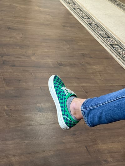 A person's left foot, clad in a green and blue checkered Vans slip-on shoe and a gray sock, rests on a dark brown wooden floor. The person is wearing blue jeans. To the right of the foot, a carpet runner with an ornamental border is visible on the floor. The scene is indoors, on a wooden floor with visible wood grain and plank lines. The carpet runner has a light beige background with a repeating geometric pattern in darker colors. There is no information in the image to suggest a specific location in Myronivka, Ukraine.