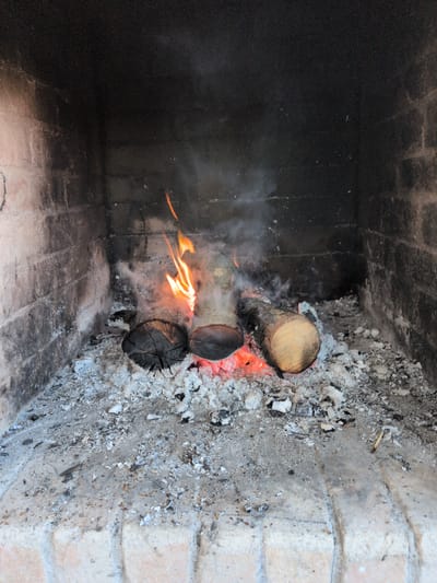 The image depicts a fireplace in Нежинка, Russia. Inside the brick fireplace, three logs are burning, with visible flames and smoke. Ashes surround the logs, covering parts of the brick structure. The left side of the fireplace is darker, contrasting with the ashy and brick-lined right side. There are no people visible in the image.