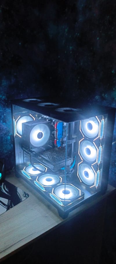 The media file displays a computer tower with illuminated fans. The computer is situated on a wooden surface, likely a desk, which is placed in front of a dark background featuring a cosmic or starry pattern. The computer itself is a black, rectangular tower with a glass side panel, allowing the internal components and the glowing fans to be visible.
There are multiple fans inside the computer, all emitting a bright white light with blue accents. Three large fans are visible on the front of the tower, and another fan is mounted on the left side, near the top. Inside the tower, other components such as RAM sticks (one blue and one red) and a graphics card are discernible. The graphics card has "GEFORCE RTX" and "INFINITI" printed on it.
The lighting from the computer fans creates a glowing effect around the device, illuminating the immediate surroundings with a cool, bright light that contrasts with the darker, starry backdrop. The overall impression is one of a modern, high-performance gaming or work setup, designed with aesthetic appeal. There are no people visible in the media file. The scene appears to be indoors, given the controlled lighting and the presence of a computer setup. The time of day and weather are not discernible. No specific location cues from Никольское, Russia, are present within the image itself.