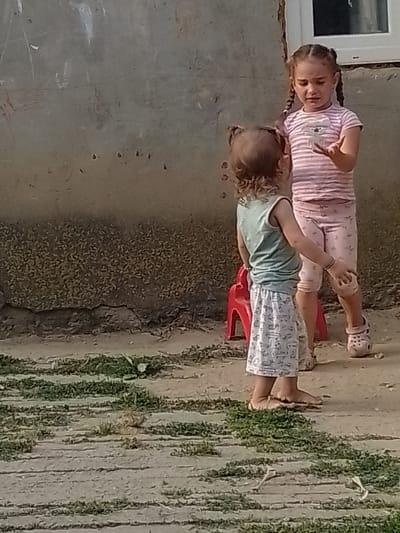Two young girls are standing in front of a grey, textured wall in an outdoor setting. The girl on the right, with braided hair, wears a pink and white striped t-shirt and pink patterned shorts. She is gesturing with her right hand, palm facing upwards, and appears to be speaking or explaining something. The girl on the left, with curly hair, wears a teal sleeveless top and a white skirt with a pattern of small figures. She is facing away from the camera, towards the other girl. A small red plastic chair is visible behind her. The ground in front of them is a mixture of dirt and patchy grass, with some uneven paving stones. The wall behind them has some faint white markings and appears weathered. The overall scene suggests a casual, everyday moment outdoors.