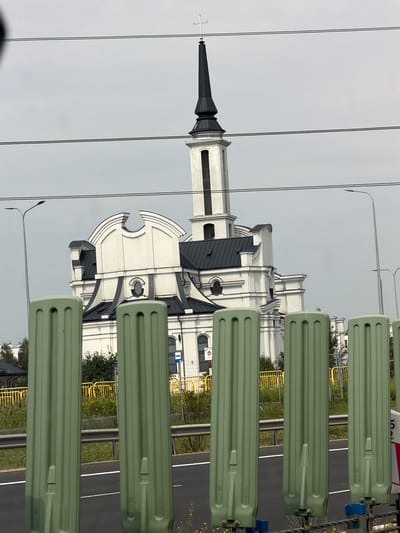 The image shows a white church with a black steeple and a cross on top, located in Dybów Kolonia, Poland. In the foreground, there are several green, vertical barriers. The church appears to be in the background, behind a road and some vegetation. There are power lines running across the top of the image. The sky is overcast.