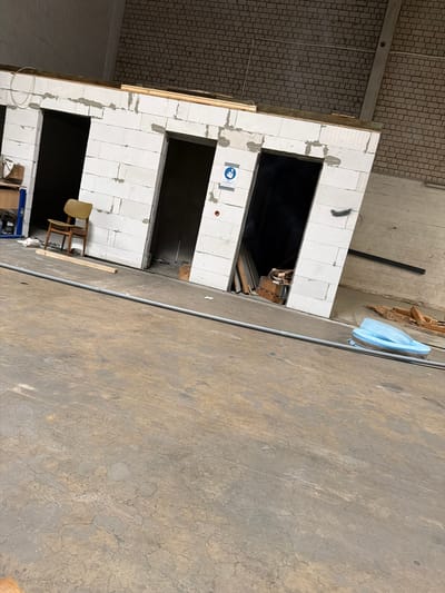 The image depicts an unfinished interior space, likely a warehouse or construction site, in Osnabrück, Germany. The primary focus is a structure built from white concrete blocks, featuring three doorways. The building appears to be in a state of construction or renovation, with exposed wooden beams at the top and loose construction materials scattered around. To the left, a wooden chair sits near a shelf unit with various items. A blue plastic object, resembling a large flip-flop or molded seating, is visible on the floor towards the right. The floor is a concrete surface showing signs of wear and tear. The background consists of brick walls and a pillar. The lighting is artificial, suggesting an indoor environment.
