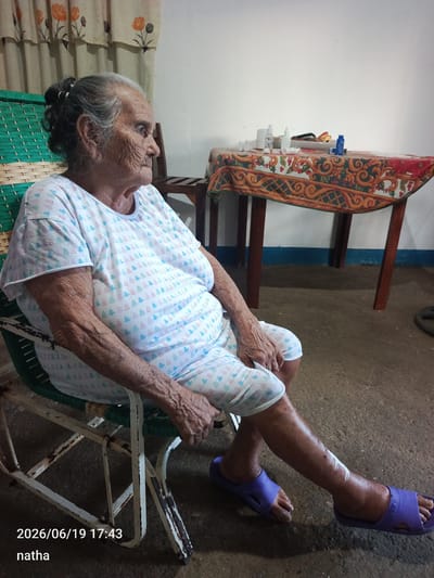 The image depicts an elderly woman seated in a rocking chair. She is wearing a light-colored pajama set with a pattern of small trees. Her right leg, which is bandaged on the lower part, is extended forward. She is wearing purple slip-on sandals. Her hair is pulled back in a bun. Behind her, a table is covered with a patterned cloth, and on the table are several bottles and objects. The environment appears to be indoors, with a tiled floor and a patterned curtain in the background. The date and time stamp "2026/06/19 17:43" and the name "natha" are overlaid on the image. The location is identified as El Chaparral, Venezuela.