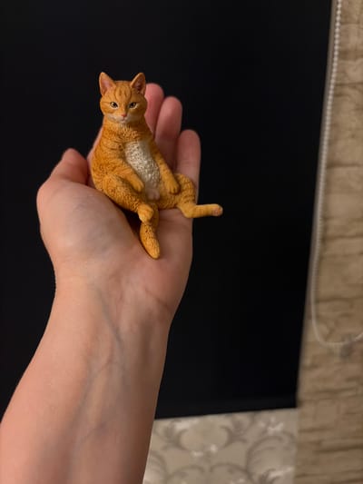 A small, orange, resin cat figurine is held in the palm of a person's hand. The cat is sitting with its legs crossed and its body tilted slightly to the left. The background is divided into two sections: the left side is a solid black color, and the right side shows a textured, beige wall with a white cord hanging vertically. The cat's fur is detailed, and its eyes are a piercing yellow. The hand holding the cat is fair-skinned and appears to be resting on a surface that is out of focus. The lighting in the scene is dim, creating shadows on the cat and hand. The scene does not appear to be located in the city of Härryda, Sweden.