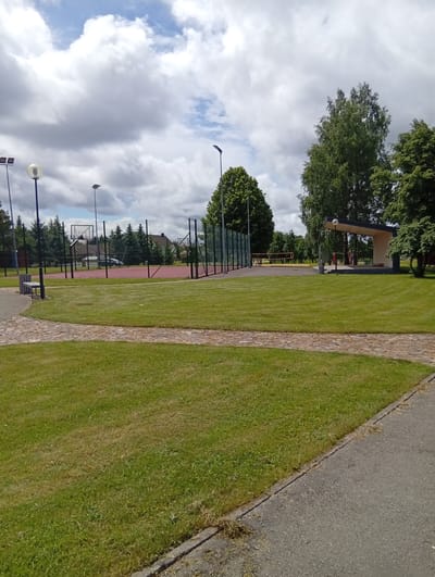This is an outdoor, eye-level, medium shot image of a park area in Šaukėnai, Lithuania, on a cloudy day. In the foreground, a paved pathway curves from the right side of the frame towards the left, bordered by a well-maintained green lawn. Beyond the lawn, a red-surfaced sports court, likely for basketball or volleyball, is visible, enclosed by a dark metal fence. Several tall, slender light poles are scattered throughout the park. In the background, lush green trees and some residential buildings can be seen. To the right, a covered open-air stage or pavilion with a light-colored roof stands amongst the trees. A lone bicycle is parked near the fence of the sports court. There are no people in the frame.