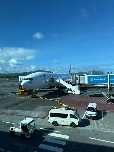 The image displays an active airport tarmac scene at Sir Seewoosagur Ramgoolam International Airport (MRU) in Plaine Magnien, Mauritius.

A white Emirates Boeing 777-300ER aircraft is centrally positioned, parked at a gate. Its nose is pointed towards the left, and its right side is illuminated by direct sunlight. A jet bridge, prominently branded with "Standard Bank" and "Africa," is connected to the aircraft's forward left passenger door. Adjacent to the jet bridge, a mobile passenger stair is deployed. A red pushback tug is visible beneath the aircraft's nose gear, with yellow chocks placed at the wheels. Fueling equipment, including yellow hoses, extends from the ground to the aircraft's right wing.

On the asphalt tarmac in the foreground, two white vehicles are parked. To the right, a white passenger van marked "AIR MAURITIUS" and "CL 10" is stationary. To its right, a smaller white utility vehicle is parked. Further left on the tarmac, a white ground support vehicle, possibly a baggage tug or tow tractor, with "AE90" visible on its side, is positioned. A person in a high-visibility vest is partially visible inside this vehicle. The tarmac features various markings, including yellow lines for aircraft parking, red striped no-go areas, and white pedestrian crosswalk-like lines.

The background reveals a clear blue sky with scattered white cumulus clouds. Distant green, forested mountains rise along the horizon, characteristic of an island environment. The overall scene indicates fair weather with bright sunlight. A faint, light-colored ring is visible in the upper sky, likely a lens artifact.