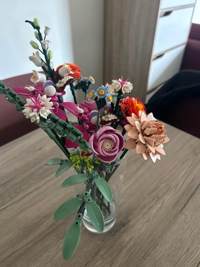 A bouquet of artificial flowers, constructed from LEGO bricks, is presented in a clear, cylindrical glass vase. The vase rests on a light-colored wooden table featuring a visible grain pattern. The bouquet comprises multiple distinct flower types and colors, including white bell-shaped blossoms with green buds, small blue flowers with yellow centers, a prominent dark pink spiral-centered flower, a layered peach-colored dahlia-like bloom, white spiky flowers with pink accents, and orange tulip-shaped elements. Green LEGO rods form the stems, which are augmented with light green leaf pieces and clusters of foliage. The background is out of focus, revealing a reddish-brown upholstered object to the left and a light wooden cabinet with white-fronted drawers positioned against a plain, light-colored wall to the right. No individuals or actions are depicted. The image does not contain any discernible visual information to identify the specific location as the city of Louviers, France.