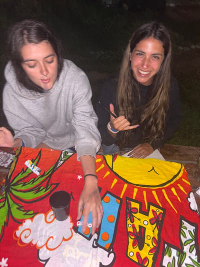 Two young women are captured in a casual setting. One, wearing a dark top with long hair, is smiling broadly and flashing a hang loose sign. The other, in a grey sweatshirt, looks down, seemingly focused on the table. A colorful tablecloth with a sun and tropical motifs covers the surface, where a lighter, a container, and cards are visible. The scene appears to be at a party or casual gathering.