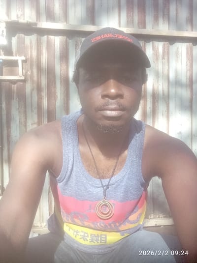 A person wearing a gray tank top with red, orange, and yellow stripes sits in front of a corrugated metal structure. They also wear a black cap with red lettering and a necklace with a circular pendant. The date "2026/2/2 09:24" is visible in the lower right corner of the image. The person's face is partially shaded by the cap, but their features are visible, including a mustache and beard. The overall lighting suggests it's a sunny day.