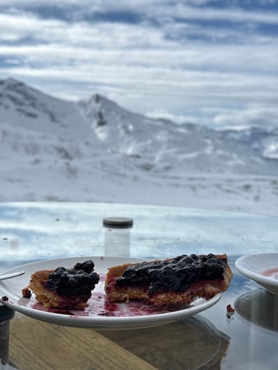 The scene depicts an outdoor setting in Saint-Martin-de-Belleville, France, with a prominent snow-covered alpine mountain range visible in the background under a partly cloudy sky. In the foreground, two partially consumed pieces of a pastry, likely a tart with a golden-brown crust and a dark, berry-based topping (consistent with blueberries or similar fruit), are centered on a round white ceramic plate. Reddish fruit juice stains are visible on the plate. A silver-colored utensil handle extends from the left edge of the plate. To the right of the plate, a small white saucer contains reddish liquid residue. Behind the plate, a clear glass bottle with a dark, possibly metallic, screw-top lid stands upright on a reflective surface, which appears to be a glass table. The table's surface shows reflections of the environment, and a wooden texture is discernible beneath it. No individuals are visible.
