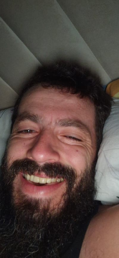 The image shows a close-up, frontal view of a man with a dark beard and hair, lying down and smiling with his eyes partially closed. He is looking towards the camera. The background is out of focus and appears to be bedding, with a textured, possibly quilted surface. A portion of a pillow is visible on the right side of the frame. The lighting suggests an indoor environment. There is no visible indication of the city of Wattenwil, Switzerland.