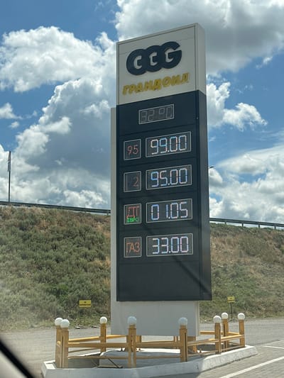 The image shows a gas price display at a CCC Grandoil gas station in Nizhniy Chiryurt, Russia. The sky is blue with white clouds. The gas price sign is tall and black with digital displays. The prices shown are: 32.90, 99.00, 95.00, 01.05, and 33.00. There is a small yellow sign in the background that reads "НЕ ПАРКОВАТЬСЯ", meaning "NO PARKING". The ground is paved with asphalt and a low yellow fence with white spherical lights encloses the base of the sign. A grassy hill is visible in the background.