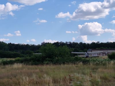 This is a medium-shot photograph of an outdoor scene with a biplane in a field. The sky is a bright blue with scattered white cumulus clouds. In the foreground, tall, dry grasses dominate the view, suggesting a summery or autumnal setting. Behind the grass, a dense line of dark green trees forms a treeline, with some lighter green bushes in front of it.
The main subject, a white biplane with blue accents, is parked on a grassy area in the middle ground, to the right of center. The plane appears to be a vintage model, with two wings stacked vertically. It has a propeller at the front and a visible cockpit where a person in a blue shirt can be seen sitting. The plane is positioned in front of a long, low building with a brown roof and weathered, light-colored walls, which appears to be a hangar or storage structure.
The scene is bathed in natural light, suggesting it is daytime, likely late morning or afternoon, given the brightness of the sky and the shadows cast by the plane. The overall impression is one of a quiet, rural setting, possibly an airfield or a private property with an aircraft. The weather appears to be clear and pleasant. No text is visible in the image.