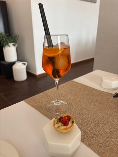 In Carboneras, Spain, on a table set with a white tablecloth and a brown burlap placemat, sits a tall, narrow glass filled with an orange aperitif and ice. Two black straws extend from the glass. In front of the glass, on a white geometric base, is a small tartlet topped with red berries, other small red and black garnishes, and a green leaf. In the background, a wooden floor is visible, along with white ceramic containers and a green plant in a white pot against a white wall. The scene appears to be indoors, likely a restaurant or bar. There are no people visible in the image.