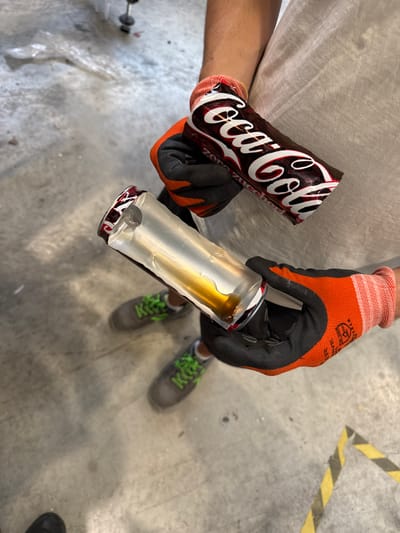 In Fontevivo, Italy, two hands, wearing orange and black protective gloves, hold two pieces of a Coca-Cola Zero Zucchero can. The top section of the can, still bearing the brand's iconic logo, is held by the left hand. The bottom section, torn open and containing a small amount of brown liquid, is held by the right hand. Both hands appear to be positioned over a concrete floor. A pair of gray sneakers with neon green laces are visible on the floor. The background is indistinct, suggesting an indoor or industrial setting.